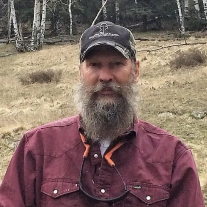 A man with long beard and a hat standing in the grass.